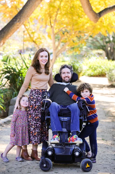 A photo of a family on a path outdoors. A man is seated in a wheelchair with a woman standing to his right and a toddler girl standing next to her. A young boy stands to the left of the man.