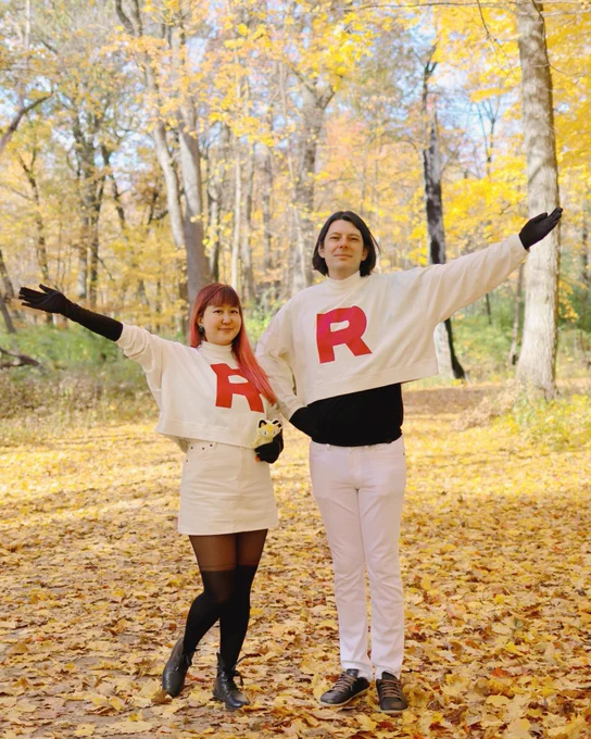 two people dressed as jessie and james from team rocket, wearing white turtlenecks that have a red R, in a forest with turning yellow leaves