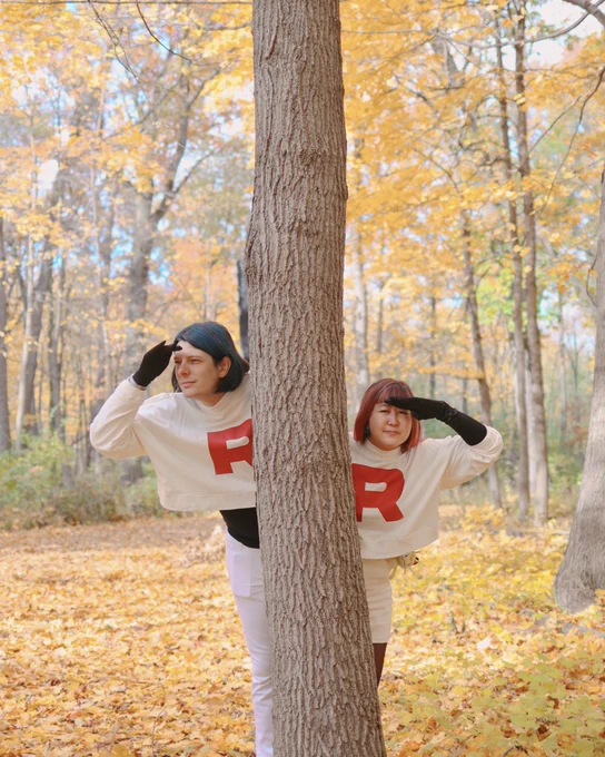 two people dressed as jessie and james from team rocket, peering from behind a tree, wearing white turtlenecks that have a red R, in a forest with turning yellow leaves