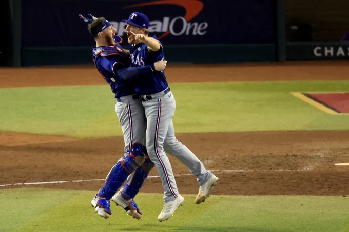 Josh Sborz and Jonah Heim jump for joy after securing the final out to clinch the World Series. They wear blue Rangers jerseys and Heim wears catcher's gear.