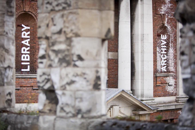 The white lettering of signs for Library and Archive are shown against a brick wall, through an out of focus stone arch