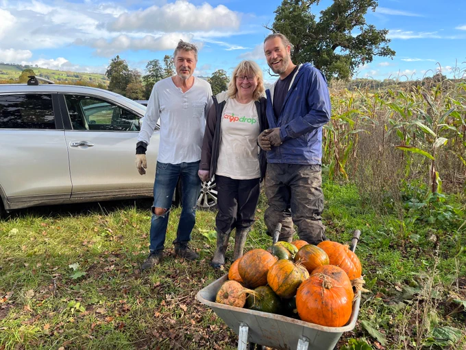 Three volunteers pose and smile for the camera beside a wheelbarrow full of gleaned pumpkins of various sizes and colours.