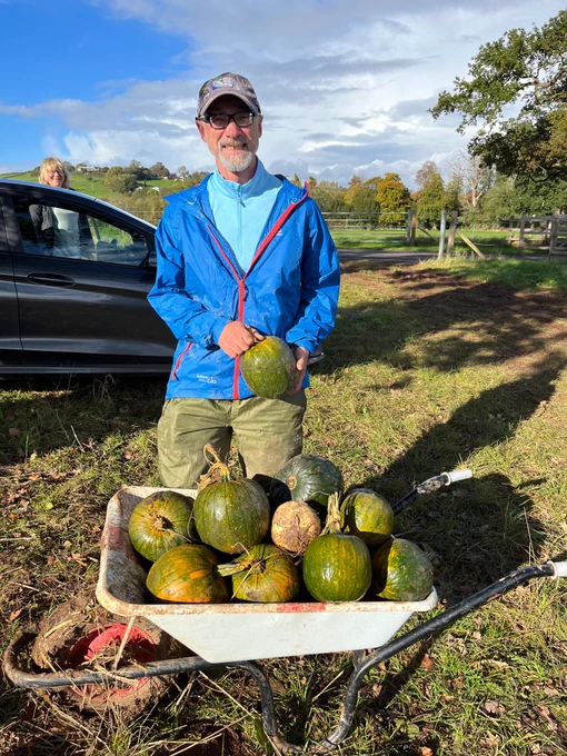 A volunteer with a bright blue jacket smiles for the camera. He is holding a pumpkin and is standing behind a wheelbarrow full of a similar variety of green-orange pumpkins.