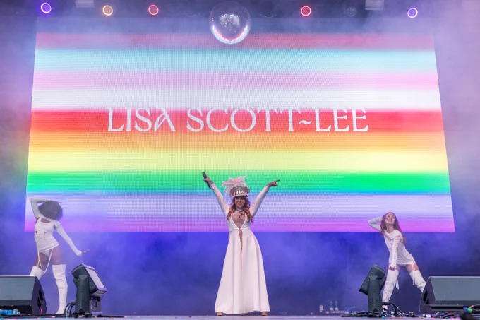 Lisa Scott-Lee and two dancers dressed in white perform on stage in front of a large screen featuring progress pride rainbow colours, reading 'Lisa Scott-Lee'.