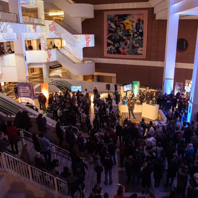 A group of people stand in the entrance hall and foyer spaces of the British Library, watching a speaker on a stage.