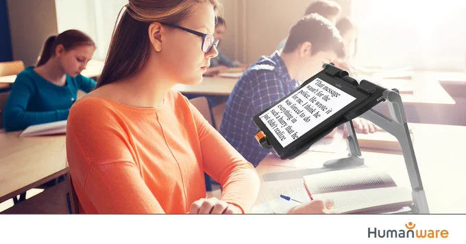 Image shows a female student sitting in a classroom. She's wearing glasses and looking at a magnifier, which is helping her to read a book below.