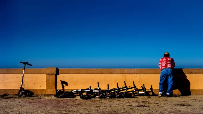 Man with his back to camera leaning on yellow block wall next to a row of abandoned scooters and looking out at blue sky.