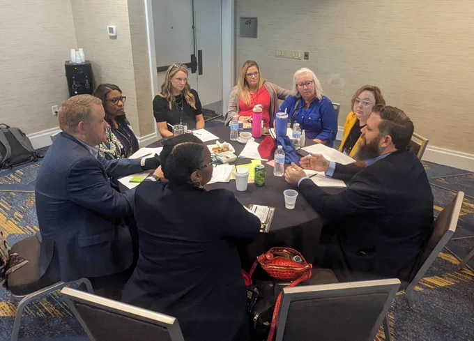 Group of diverse individuals sitting around a table with a black tablecloth and apers and notes on the table having a discusion. 