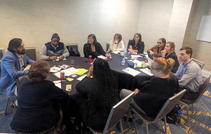 Group of diverse individuals sitting around a table with a black tablecloth and apers and notes on the table having a discusion. 