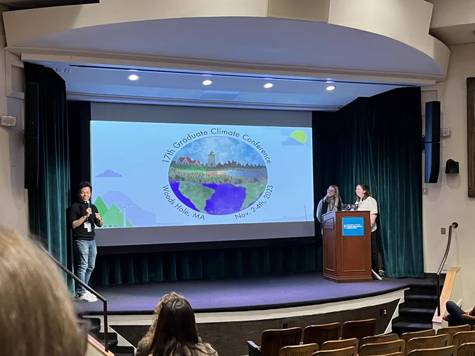 On stage, three organizers who are grad students hold microphones to welcome everyone to the Graduate Climate conference, or GCC. The projector screen shows the GCC logo with a picture of houses, trees, and a blue sky, which fades into an image of the globe with south america and africa in view.