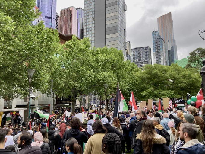 Photo taken from the back of a crowd of a few thousand people. There are Palestinian flags throughout the crowd.