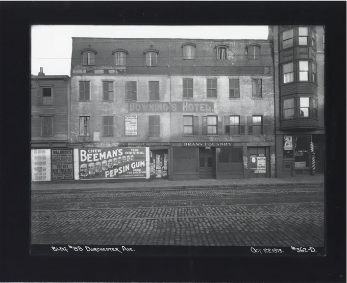 Cobblestone street with brick building with sign "Downing's Hotel" and "Brass Foundry." Large advertisement for Beeman's Pepsin Gum is plastered onto the building
