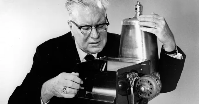 Black and white photograph of Chester Carlson looking into a xerographic device that could copy documents quickly. Carlson has short, white hair and is wearing a black suit and a pair of black-framed glasses. Carlson is embracing the device with both arms.