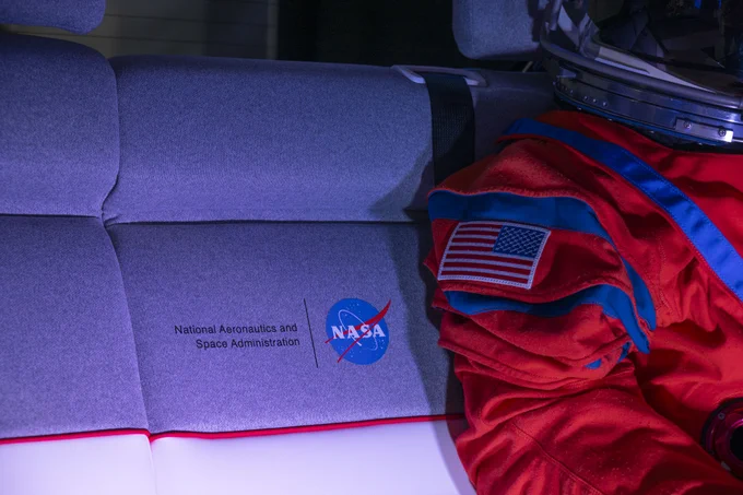 A closeup shows NASA's logo on the middle of the back seat. The seat is gray on the upper portion and white on the lower, with red piping dividing the two. The orange arm of "Campos" is in view.
Photo Credit: (NASA/Robert Markowitz)