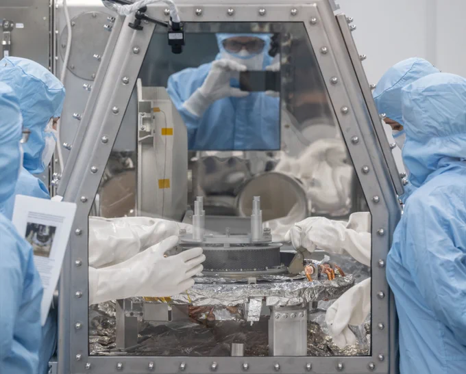 Five people in blue clean room gowns and white gloves stand around a glass and metal glove box with a circular sample head inside.