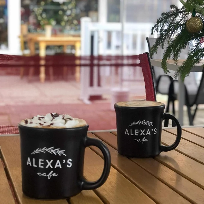 Two black coffee cups that read Alexa’s Cafe, one topped with whipped cream and chocolate drizzle, sit on a, outdoor wood table.
