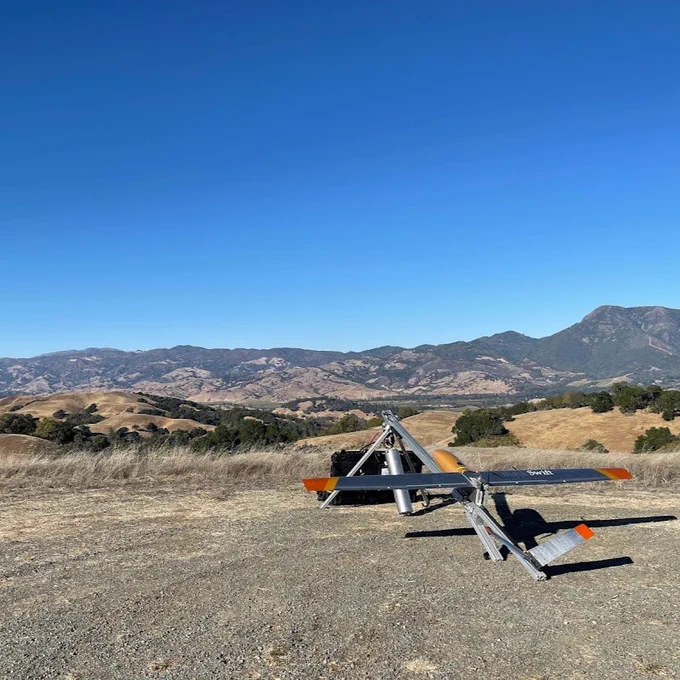 An electric uncrewed aircraft system equipped with remote sensing technology is sitting on the ground, overlooking a landscape of sloping hills covered in short, dry grass and dark green bushes. Credit: USGS