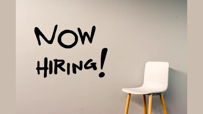 An office chair sits against a wall with the words "Now Hiring!" painted onto it.