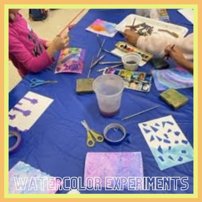 Kids work at a table covered in a blue tablecloth. There are scissors, water cups, paintbrushes, watercolor paints, rolls of tape on the table, finished and unfinished art pieces. Three kids' arms can be seen working.