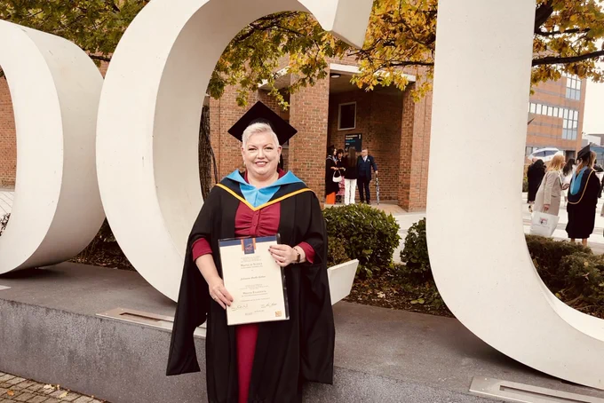 Middle age lady with short white hair wearing black masters graduand gown and cap with navy & blue sole edged in gold; she’s holding a graduate parchment in front of giant DCU letters in white.
