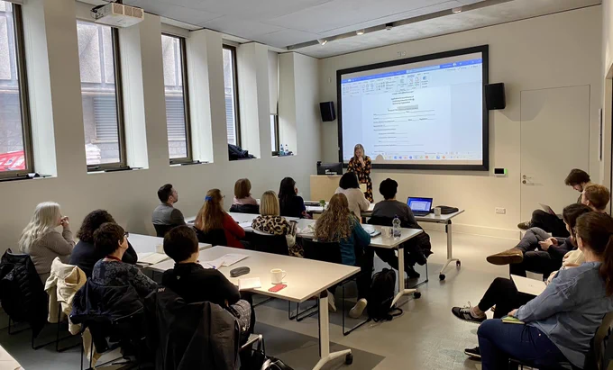 Certainly!

A classroom setting with Sophie presenting at the front beside a projected screen displaying a document. Attendees are seated at tables, listening and taking notes. The room has large windows on the left wall."