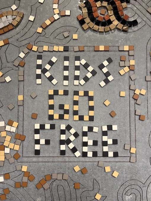 A magnetic board with mosaic tiles in brown, beige, white and black. The words ‘KIDS GO FREE’ have been created with the tiles