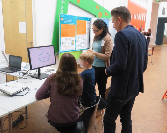 Drei Personen am Stand des IDS mit Rahaf Farag. Alle schauen auf einen Monitor. Im hintergrund zu sehen ist das Plakat zur Arbeit des IDS. Fotos: IDS