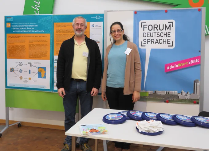 Rainer Perkuhn und Rahaf Farag stehen vor zwei Aufstellern, einer enthält Infos zum "Erforschen der Sprache mithilfe informatischer Methoden", einer trägt die Aufschrift "Forum Deutsche Sprache". Im Vordergrund ist ein Tisch zu sehen mit kleinen Geschenken für die Besuchenden, darunter Frisbees. Fotos: IDS