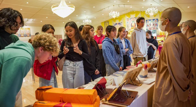 Photo of university students speaking with Buddhist nuns as they are gathered around a table holding a variety of Buddhist paraphernalia. 