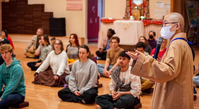 Photo of a group of university students sitting cross-legged in a buddhist prayer hall while Buddhist nun stands in front of them gesticulating.
