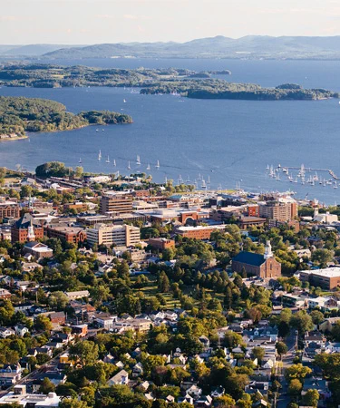 Aerial view of Burlington, Vermont, showcasing the cityscape with prominent buildings, nestled neighborhoods, and green foliage. In the background, the vast expanse of Lake Champlain stretches out, dotted with sailboats, and is bordered by rolling hills and distant mountains, the Adirondack, in New York state.