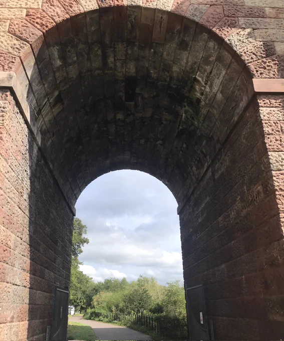 A photograph of a tall sandstone archway, through which we can see a tree lined path curving away to the left and a cloudy sky. The camera is angled up so we can see the underside of the archway curve.    

Image: © Wendy Connelly
