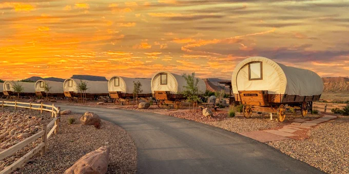 Covered wagons line up on a ranch with a sunset behind them.