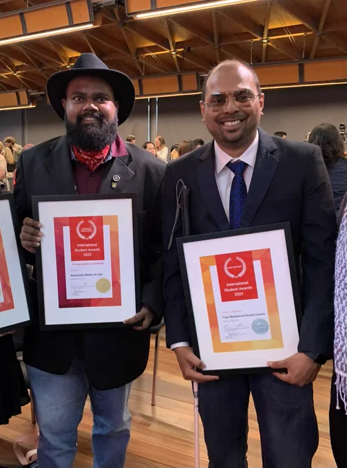 Two men stand side by side, smiling. They are both holding framed award certificates.