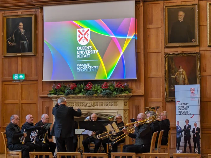 A brass band with conductor play in the wood-panelled Great Hall in Queen's. A screen above them reads, 'Queen's University Belfast Prostate Cancer Centre of Excellence'. A banner to their right reads, 'Support Protate Cancer Research at Queen's.'
Framed portraits are visible on the walls.