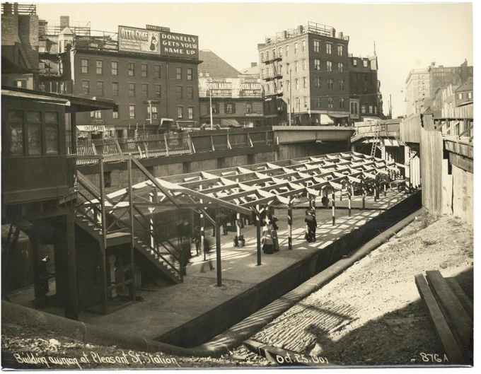 Sepia photo of outdoor transit platform with wooden frame for awning being built. Passengers stand on platform
