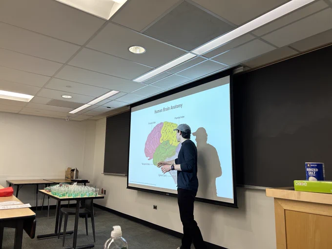 A man dressed in dark clothing and a hat stands in front of a classroom with a diagram of the human brain projected on the screen behind him.