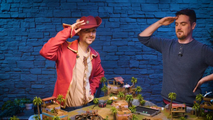 Lewis and Ben stand in the Yogscast Games Night room. The brick wall behind them in bathed in blue light. They're both saluting. Lewis wears a red militar jacket and a red pirate hat. There's a board in front of them covered in a miniature landscape; primarily palm trees and beach huts.