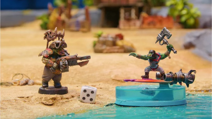 A close up shot of two miniatures and a dice. One orc stands on the beach shore while the other "floats" on a surfboard (made to look lik it's hovering) weilding an axe.