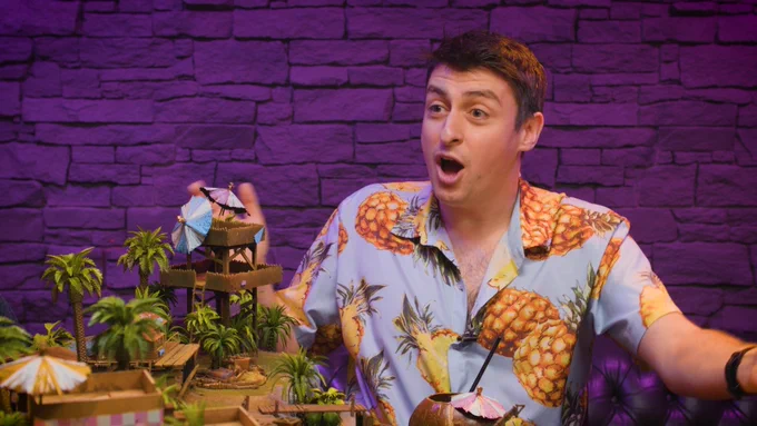 Simon Clark sits in the Yogscast Games Night room and wears a stunned expression and a tropical shirt. The brick wall behind him is bathed in pink light. There's a board in front of him covered in a miniature landscape, primarily palm trees and beach huts. 