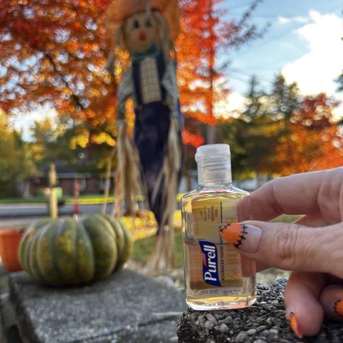 Travel-size PURELL hand sanitizer sits in front of a scarecrow and a pumpkin in a beautiful fall scene. Bottle is held by hands with nails manicured with an orange and black Halloween theme.