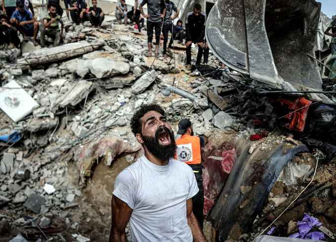 A man reacts, standing beside a huge pile of rubble following an airstrike. Workers and onlookers stand nearby and atop the rubble.