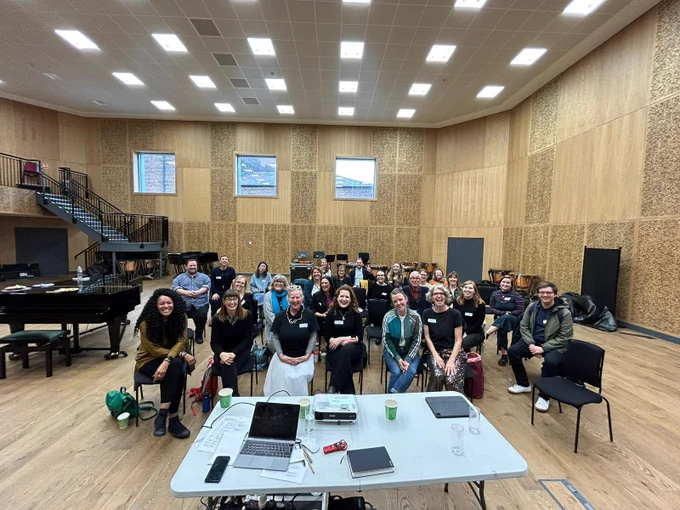 This is a wide angle photo of a group of people sitting on chairs in a large, bright, wooden panelled room. There is a table with a laptop and a projector in the foreground.
