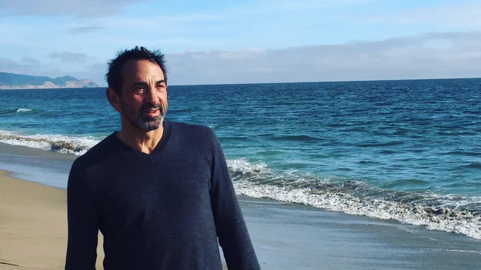 A person in a dark blue sweater walking along the shore with waves rolling onto a sandy beach behind them.