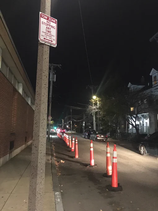 A bike lane is demarcated on a street using tall safety cones. A sign is posted on the lamppost reading “TOW ZONE; NO STOPPING ANY TIME; ASHMONT BRANCH DIVERSION; OCT 14 - OCT 29”.