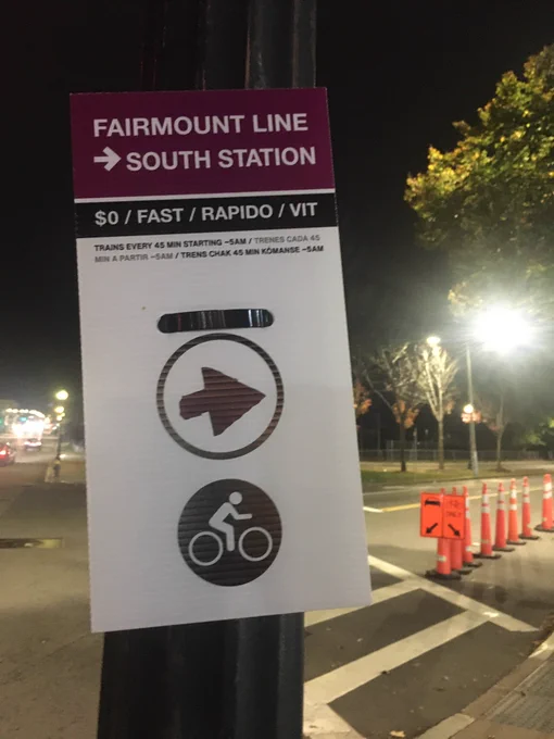 A sign posted on a traffic signal post direct bicyclists to turn right for the route to “FAIRMOUNT LINE -> SOUTH STATION”. It is noted that the train is “$0” and, in multiple languages, that service is fast with trains every 45 minutes beginning at around 5 AM.