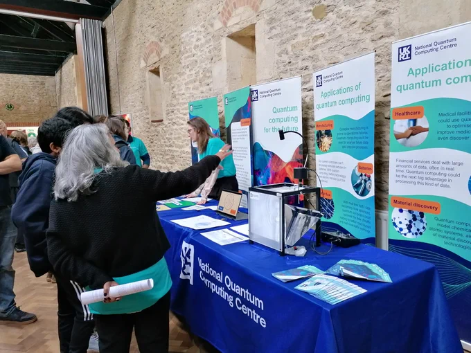 A stand at a public engagement event. There are tables with national quantum computing centre tablecloths, banners with information about quantum computing and volunteers speaking to members of the general public. There is also a ripple tank demo showing interference and wave physics.
