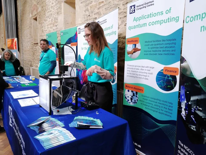A stand at a public engagement event. There are tables with national quantum computing centre tablecloths, banners with information about quantum computing and volunteers speaking to members of the general public. There is also a ripple tank demo showing interference and wave physics.