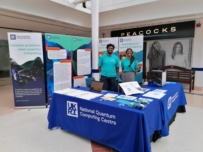 A stand at a public engagement event. There are tables with national quantum computing centre tablecloths, banners with information about quantum computing and NQCC outreach volunteers. There is also a ripple tank demo showing interference and wave physics and other quantum computing demos.