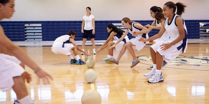 High school students in gymnasium run toward balls; coach or teacher observes in the background